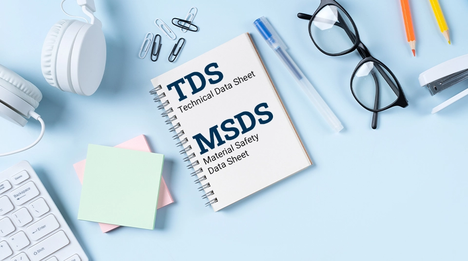 Czym różni się karta techniczna od karty charakterystyki_zdjęcie biurka z góry, biała klawiatura, białe słyuchawki karteczki, notes z nampiesm TDS i MSDS spinacze, niebieski długopis, okulary z czarnymi oprawkami, biały zszywacz, kredka żółta i pomarańczowa, wszystko lezy na błękitnym biurku