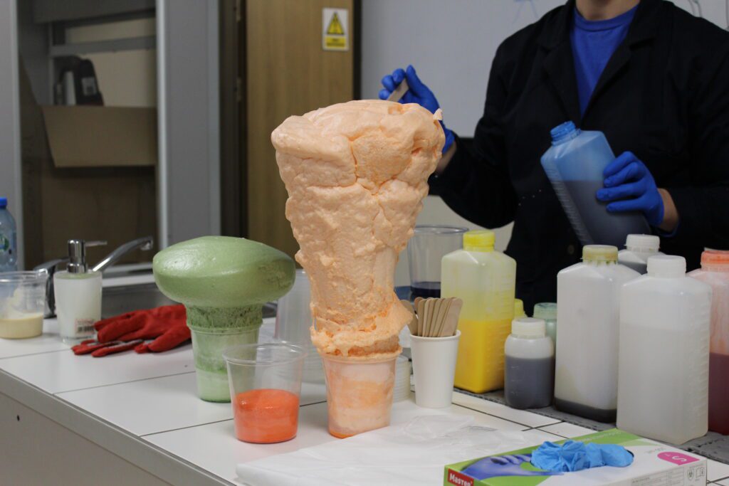 Spotkanie z Nauką: Efektowny eksperyment chemiczny, gdzie pomarańczowa i zielona piana poliuretanowa gwałtownie rośnie w plastikowych kubkach na laboratoryjnym stole. W tle widać osobę w fartuchu i niebieskich rękawiczkach trzymającą butelkę z płynem, a także różnorodne odczynniki chemiczne.