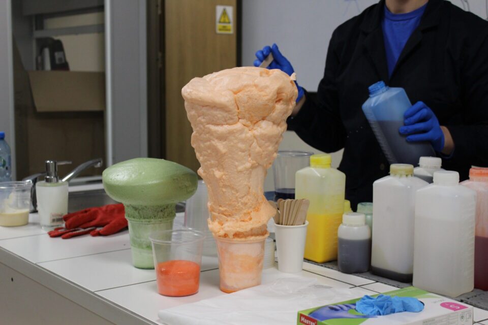 Spotkanie z Nauką: Efektowny eksperyment chemiczny, gdzie pomarańczowa i zielona piana poliuretanowa gwałtownie rośnie w plastikowych kubkach na laboratoryjnym stole. W tle widać osobę w fartuchu i niebieskich rękawiczkach trzymającą butelkę z płynem, a także różnorodne odczynniki chemiczne.