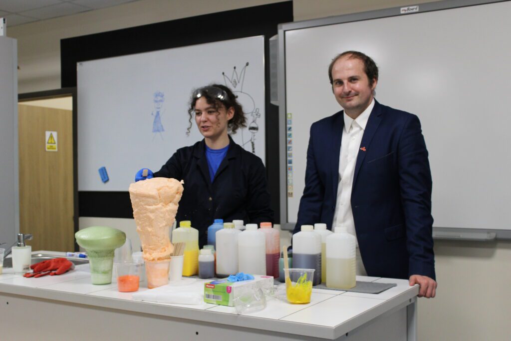 2 osoby stoją w klasie naukowej i robią eksperyment chemiczny. Kobieta jest ubrana w strój chemika, ma fartuch, rękawiczki i okulary ochronne. Mężczyzna jest w garniturze.