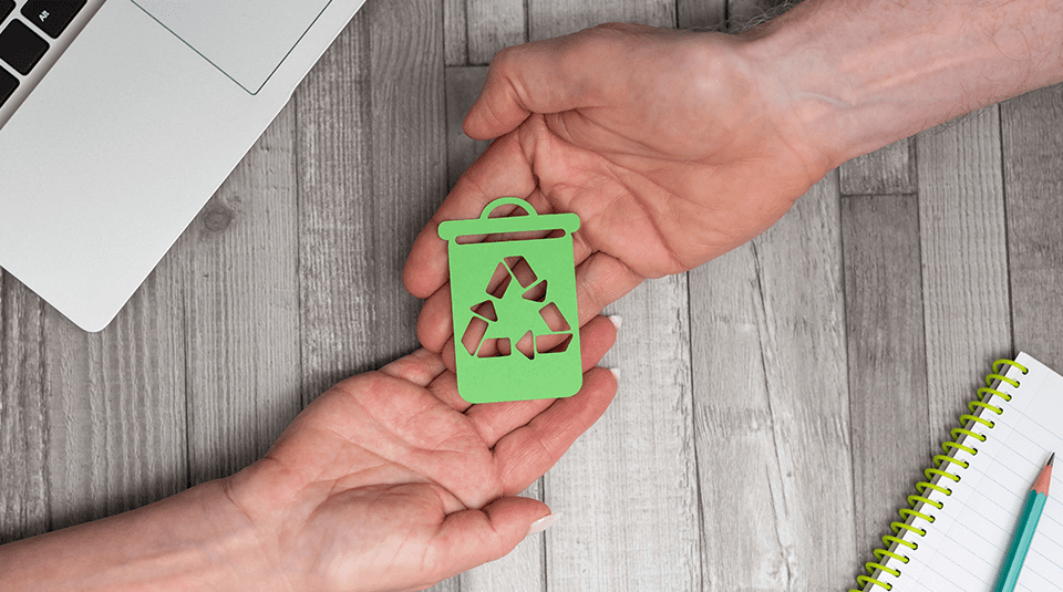 Dwie dłonie trzymające zielony papierowy symbol kosza na śmieci z logo recyklingu, umieszczone na drewnianym blacie obok laptopa i notatnika.