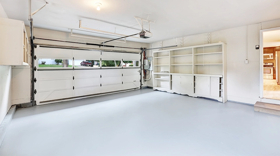 Czysty, jasny garaż z białą bramą i regałami, w którym na posadzce zastosowano wytrzymały polimocznik na zimno w jasnoszarym kolorze.